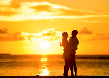 A couple on a honeymoon toasting with the sun shining over the water at dusk.