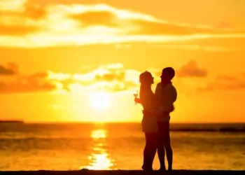 A couple on a honeymoon toasting with the sun shining over the water at dusk.