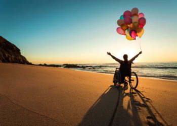 A person on a wheelchair with colorful balloons at the beach