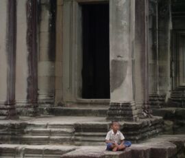 A child sits cross-legged outside a stone temple in Angkor Wat, Cambodia