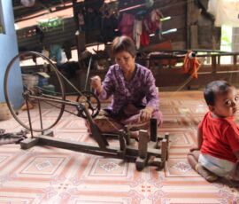 A Cambodian woman weaves traditional cloth, while her baby looks on.